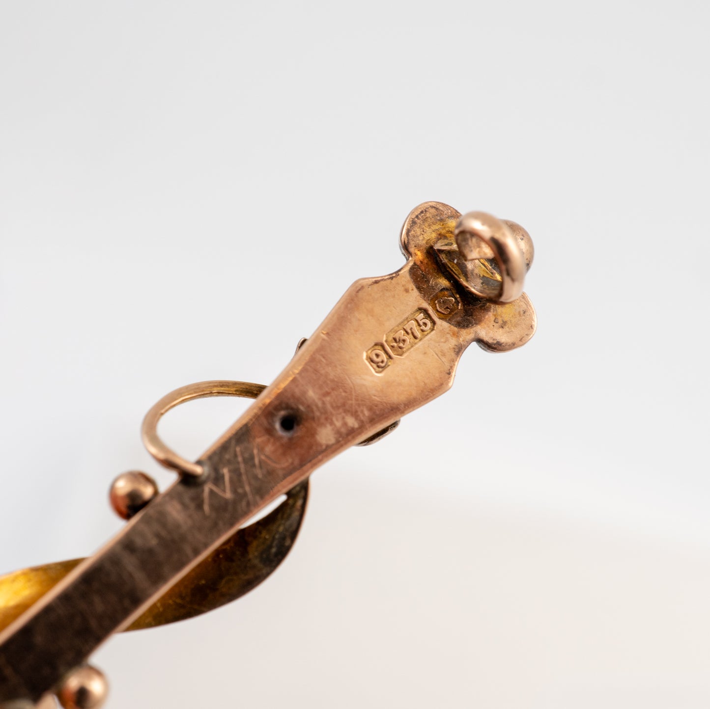 Close-up detail of a vintage gold-toned metal brooch or pin, showing engraved maker's marks and a decorative top loop, on a plain light gray background.