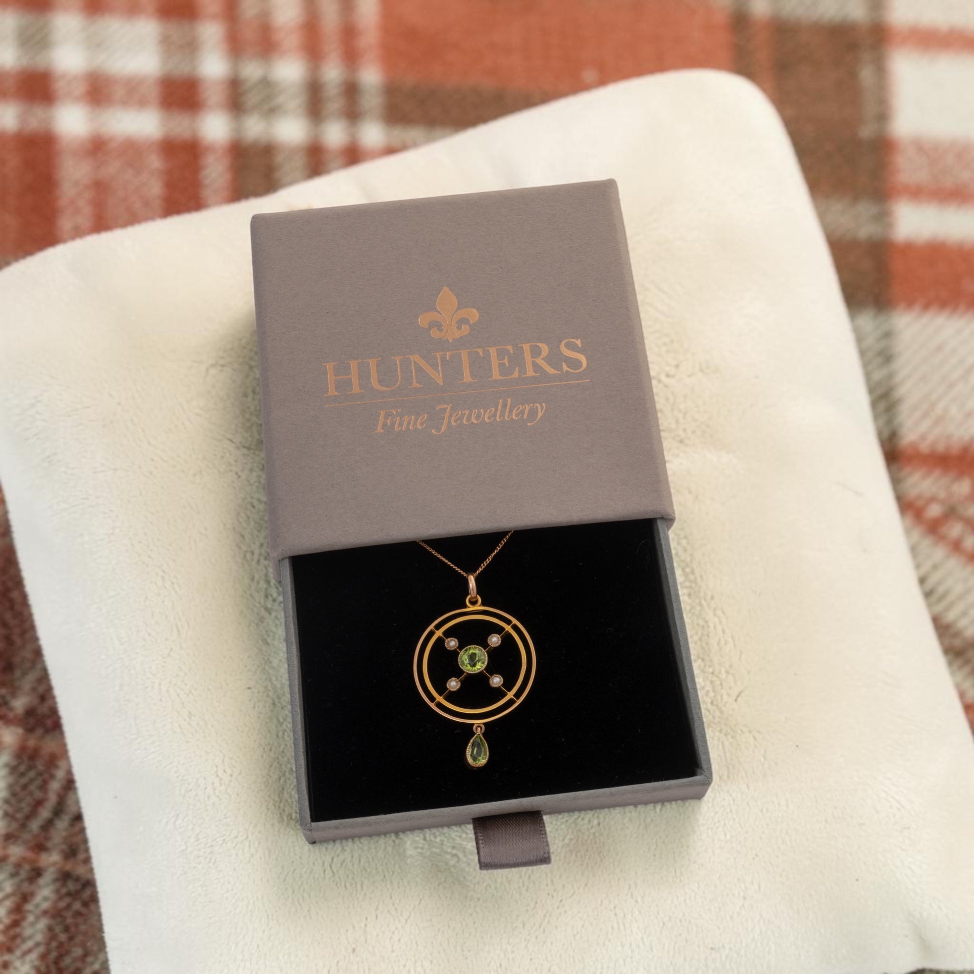 A gold pendant necklace with a green stone and small clear stones, presented in a dark gray Hunters Fine Jewellery box on a white textured cloth over a plaid blanket.