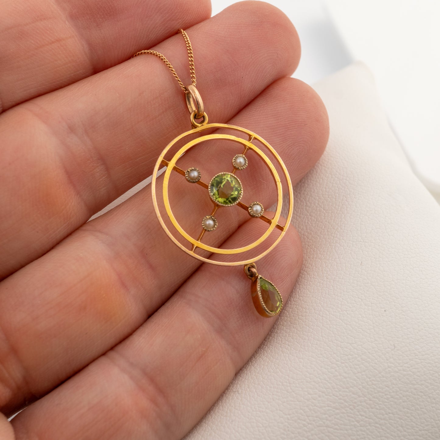 A gold pendant necklace with concentric circular rings, a central green gemstone, small pearl-like beads, and a dangling green teardrop stone, held in a person's hand against a light background.