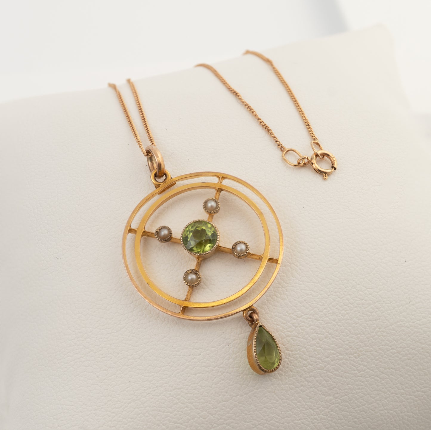 A gold pendant necklace with a circular geometric frame, a central green gemstone, small clear accent stones, and a dangling green teardrop gemstone, displayed on a white textured surface.