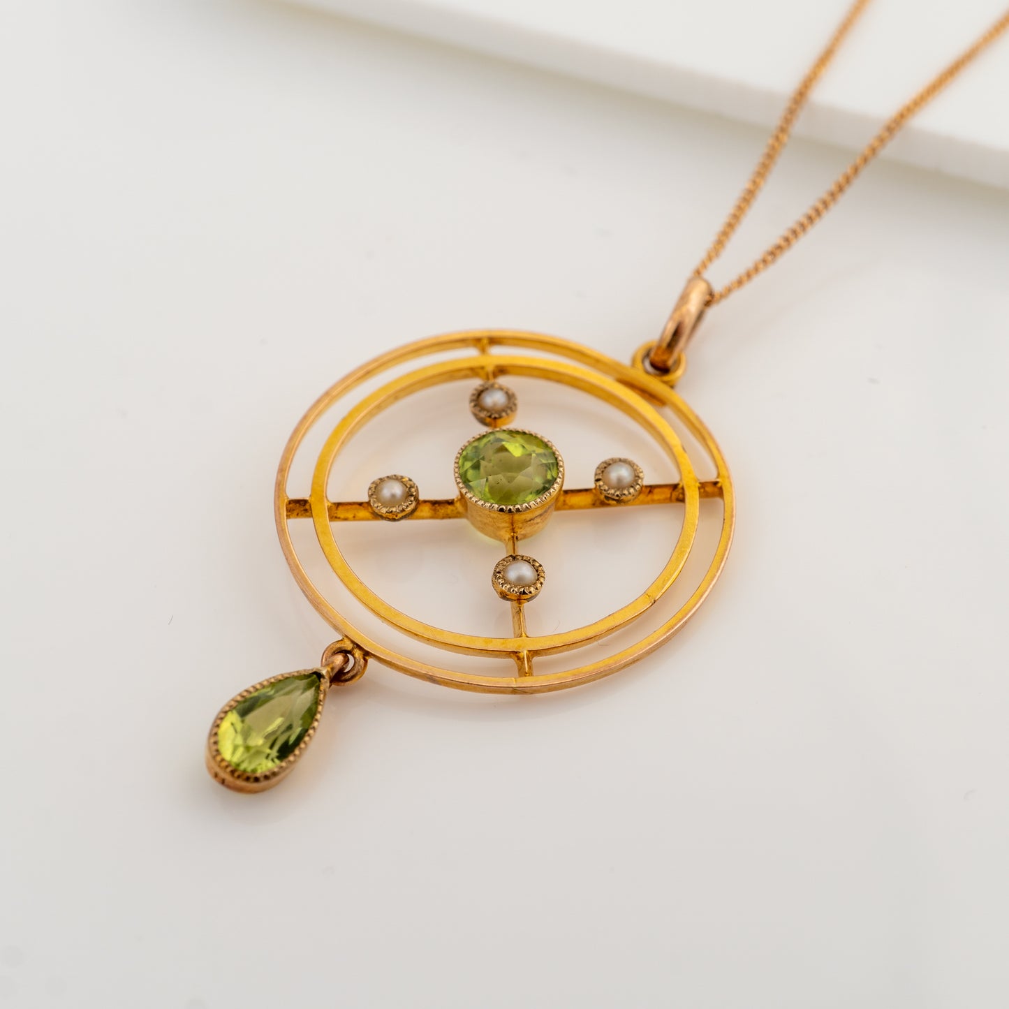 A gold pendant necklace featuring a geometric circular design with a central green gemstone, small pearl-like accents, and a dangling green teardrop gemstone, shown on a clean white background.