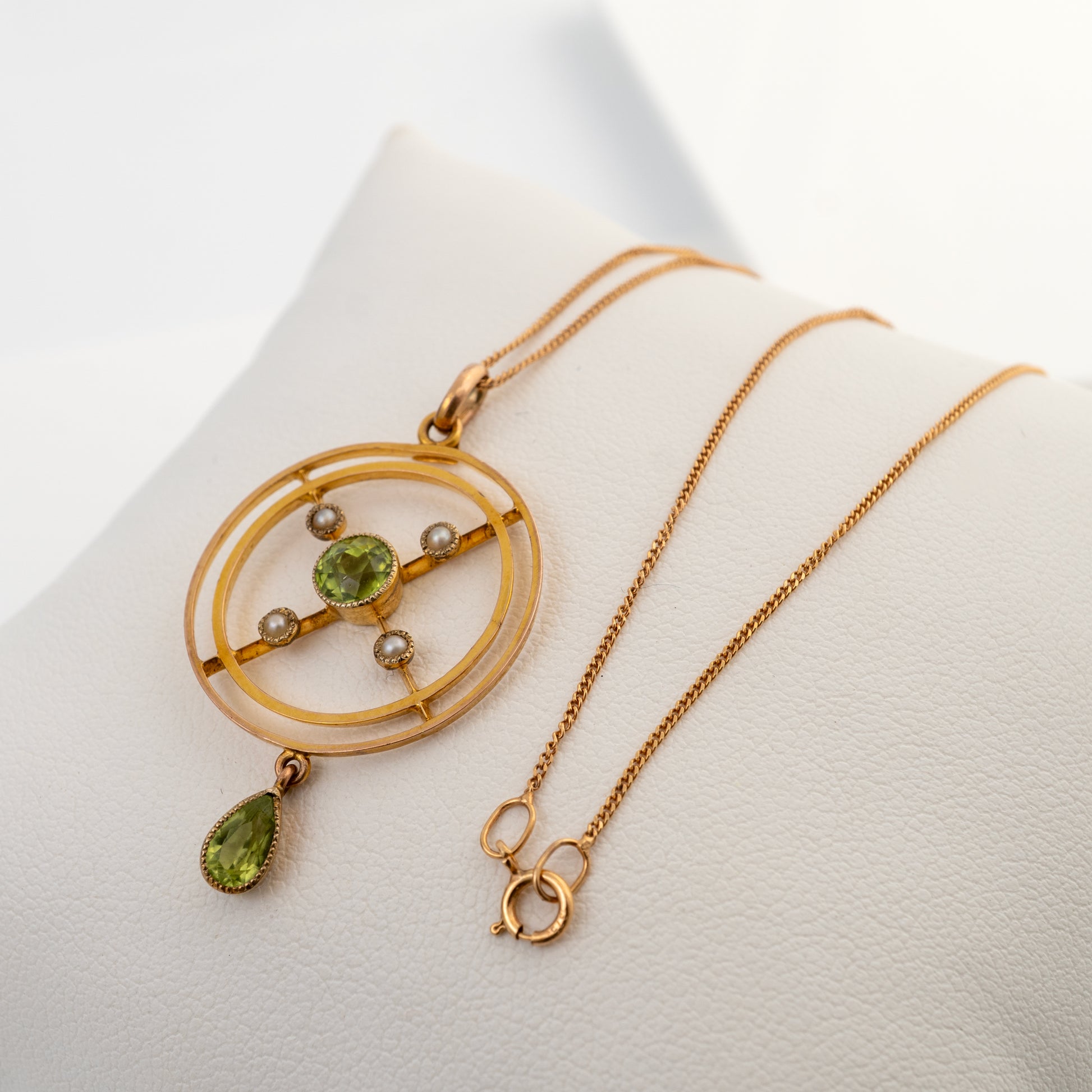 A gold pendant necklace with a geometric circular design, featuring a central green gemstone, small pearl-like beads, and a dangling green teardrop gemstone, displayed on a white jewelry stand.