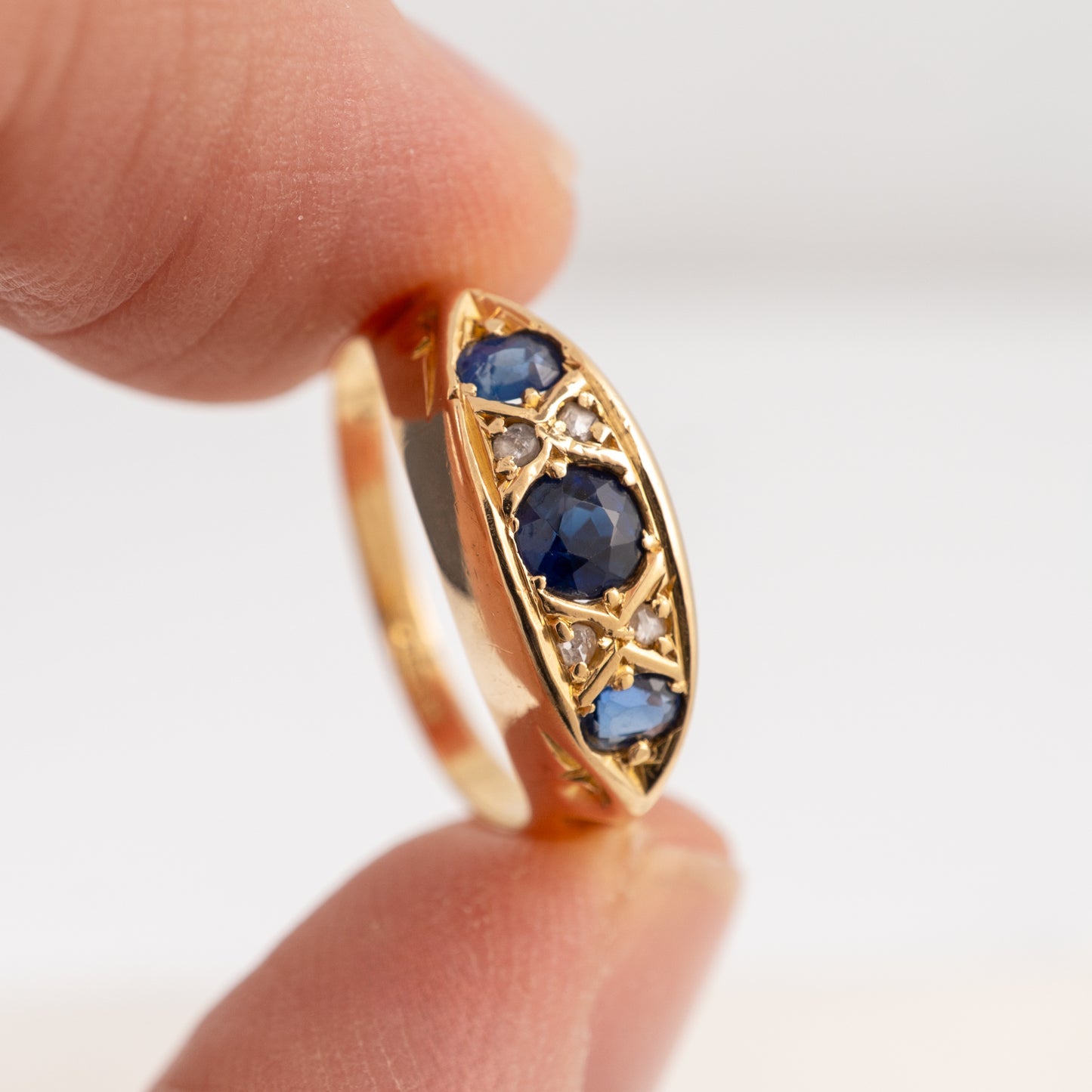 A gold ring with three blue sapphires and small clear accent stones, held between fingers against a soft white background.