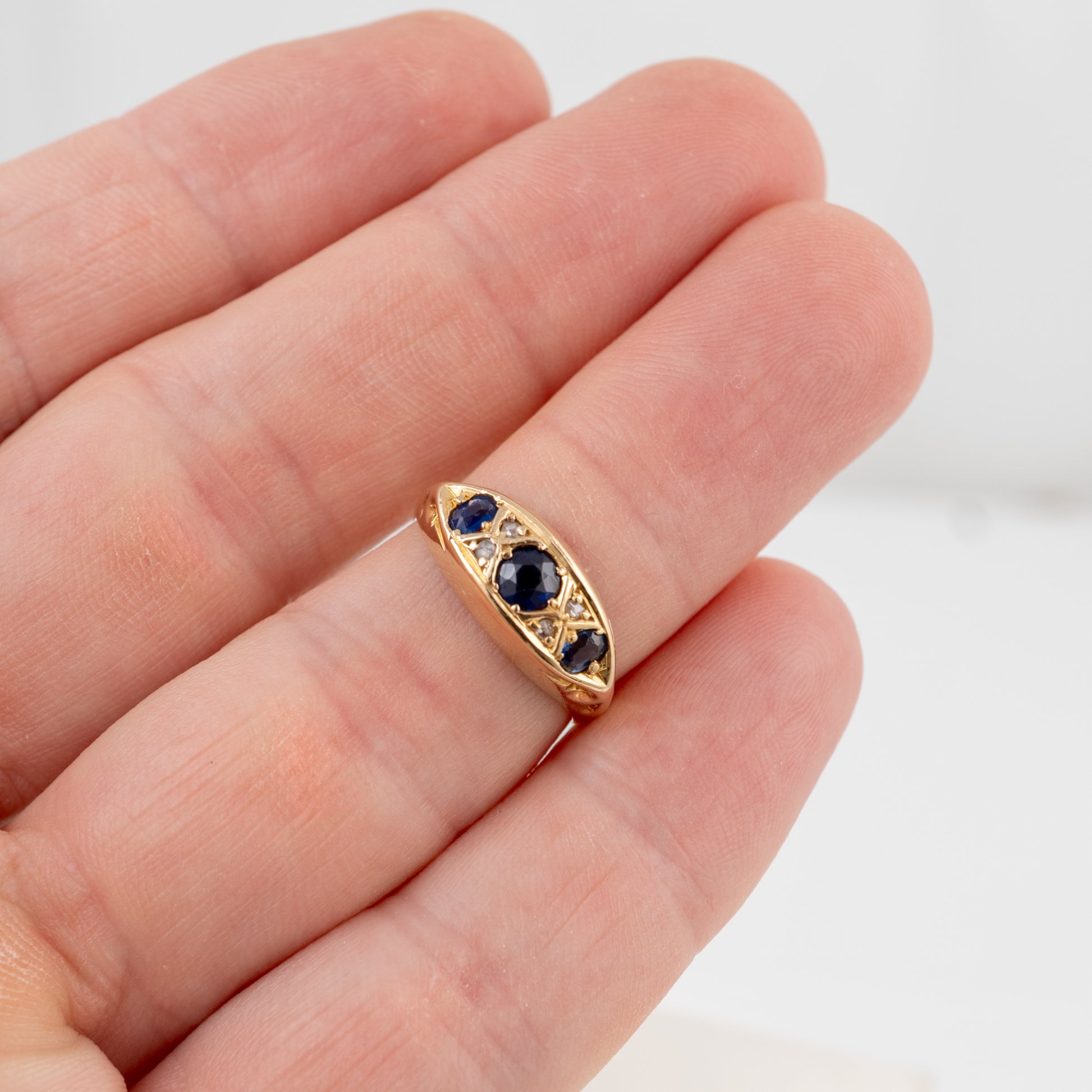 A gold ring with three dark blue oval gemstones, shown resting on a person's fingers against a white background.