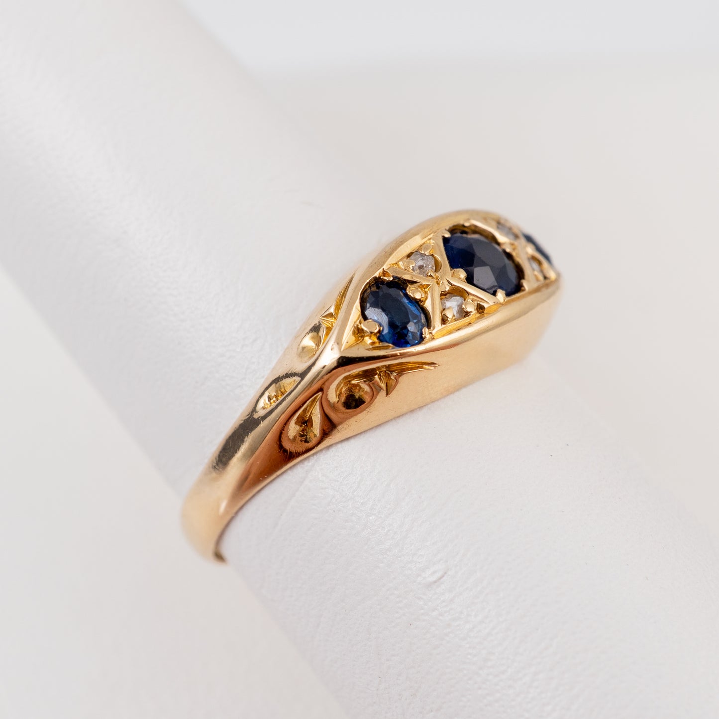 A yellow gold ring with three dark blue sapphires and small clear accent stones, displayed on a white cylindrical ring stand.