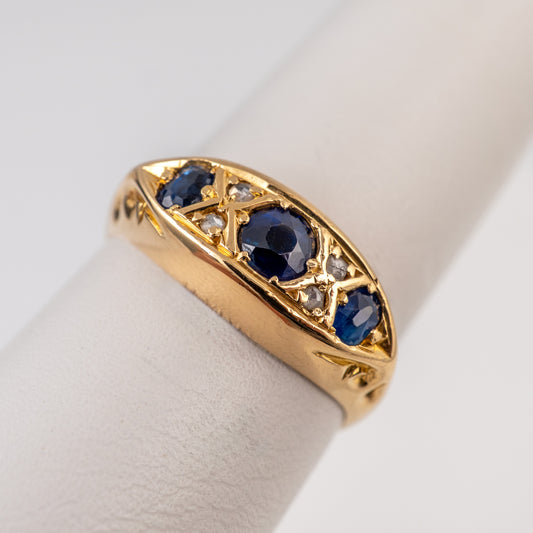 antique sapphire ring with three round blue sapphires and small clear stones, displayed on a white ring stand against a light gray background.