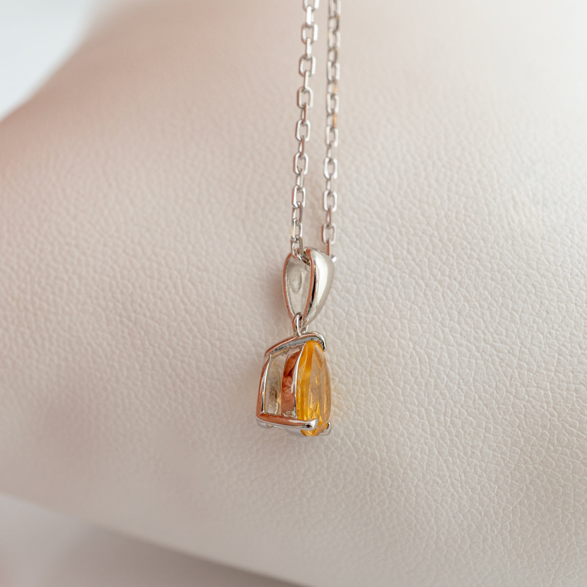 A silver pendant necklace with a faceted amber-orange gemstone, displayed on a textured cream-colored surface.