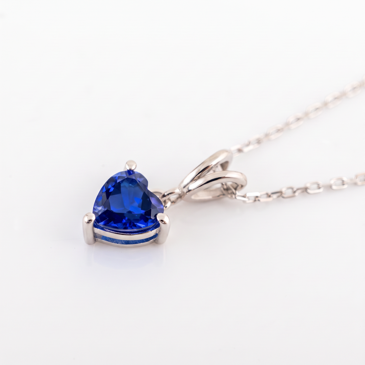 A silver pendant necklace featuring a heart-shaped blue gemstone in a prong setting, shown on a clean white background.