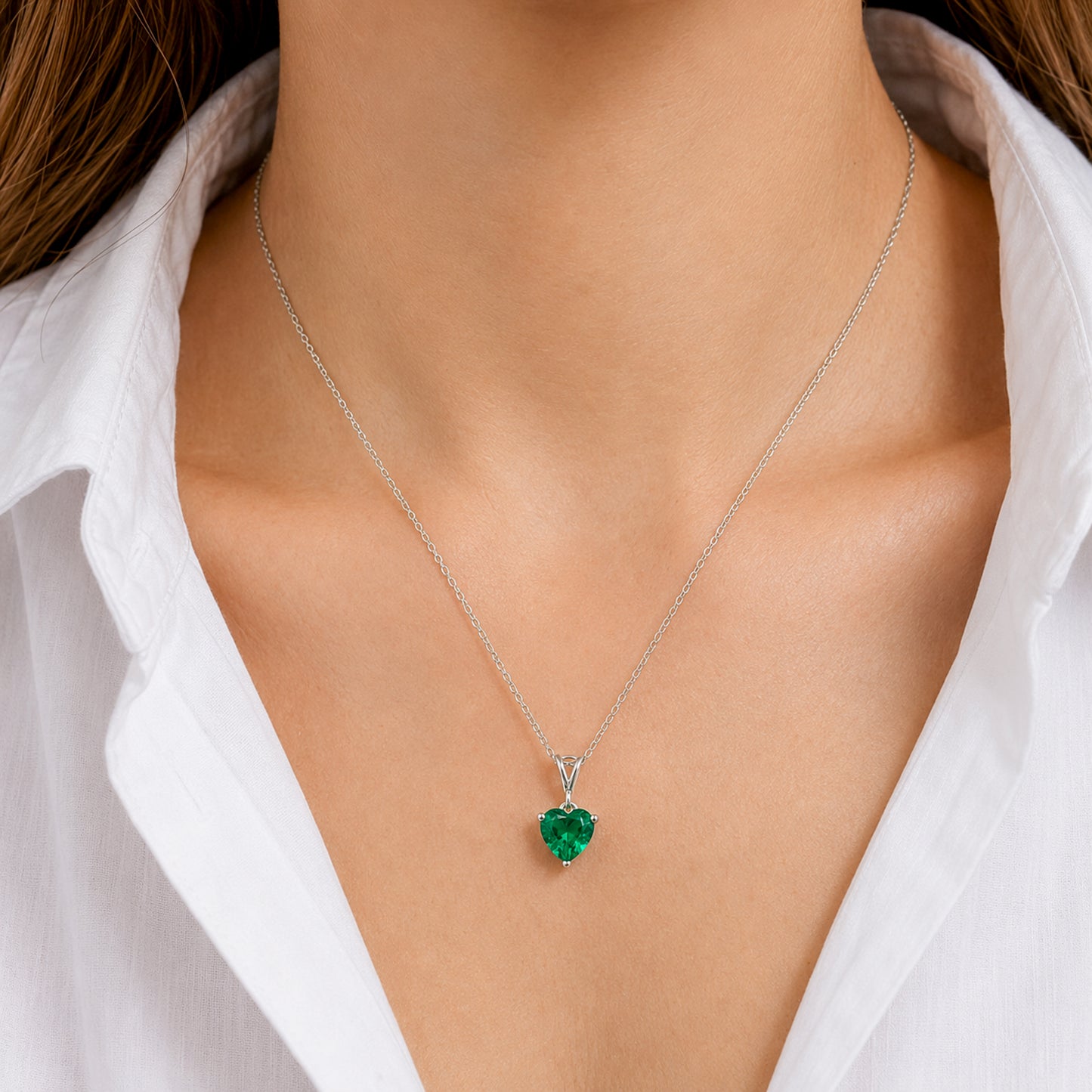 A close-up of a model wearing a delicate silver chain necklace with a small, heart-shaped green gemstone pendant, styled with a white collared shirt.