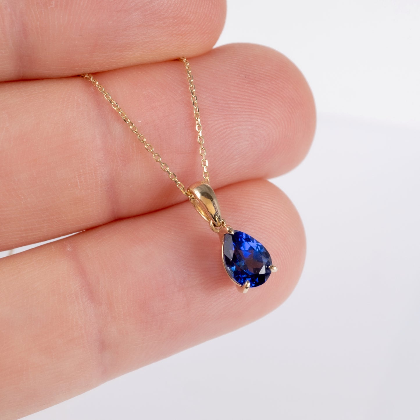 A gold chain necklace with a teardrop-shaped deep royal blue sapphire gemstone pendant, held between fingers against a plain white background.