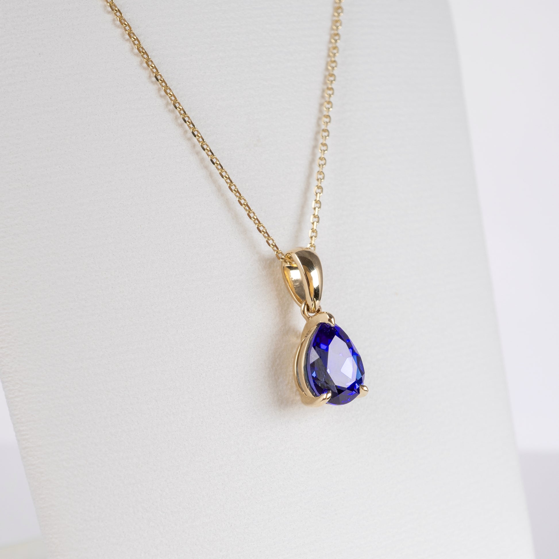 A gold chain necklace with a teardrop-shaped deep blue gemstone pendant, displayed on a white jewelry bust.