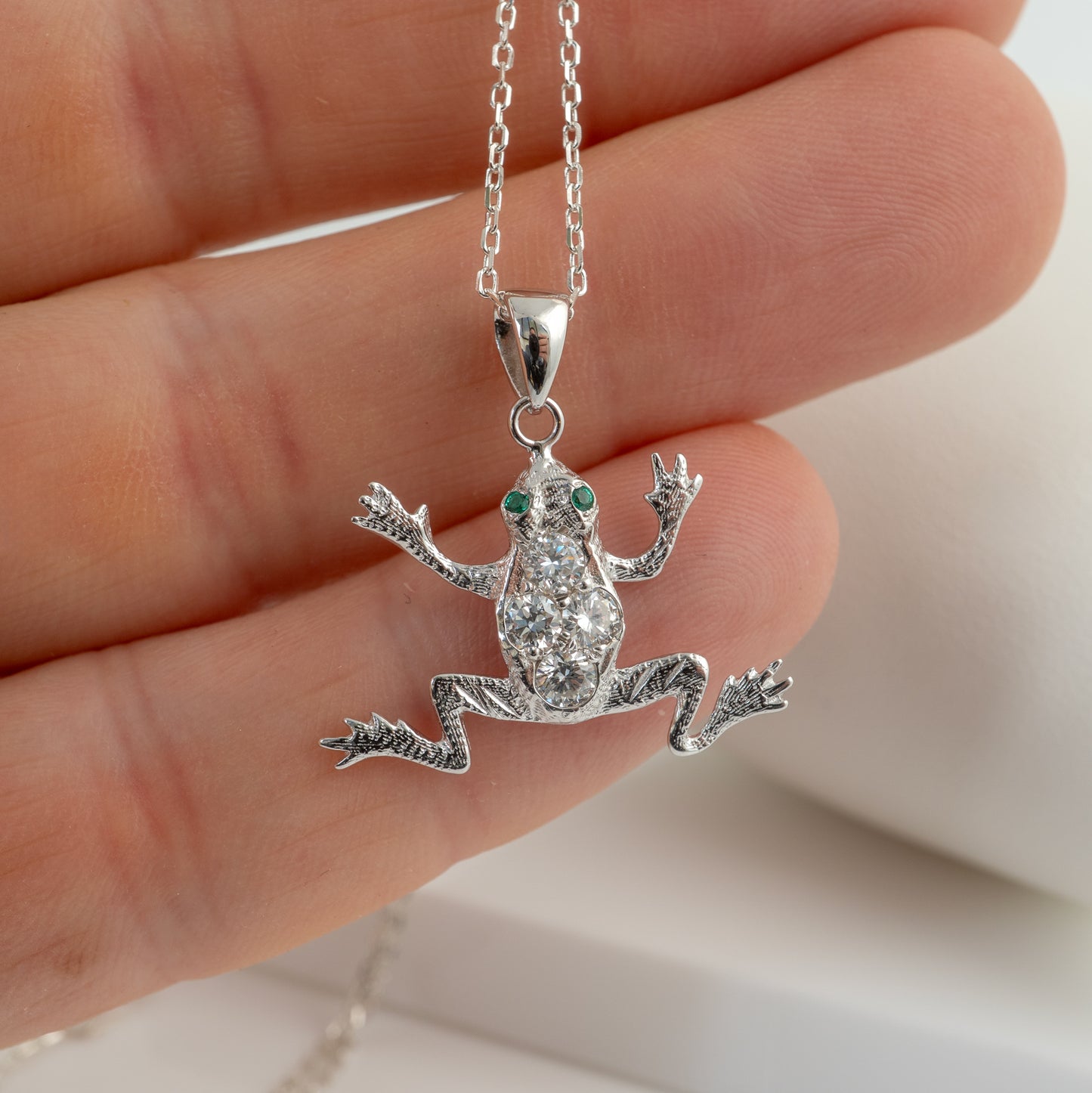 diamond frog pendant with green eyes held in a hand against a neutral background