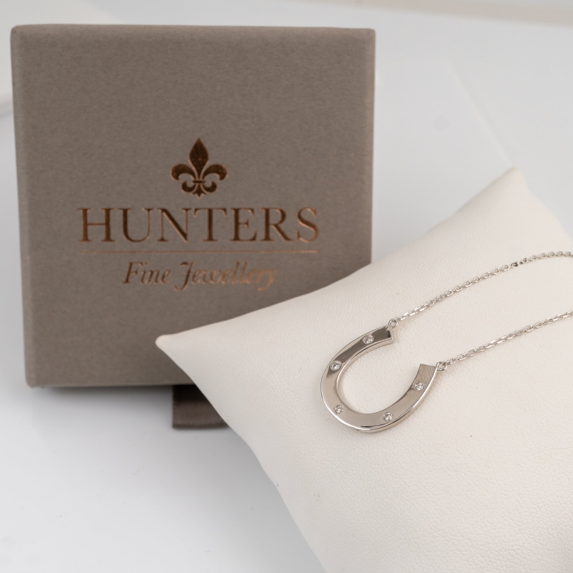A silver horseshoe pendant necklace displayed on a white cushion, with its branded taupe presentation box behind it. The box is embossed with the Hunters Fine Jewellery logo.