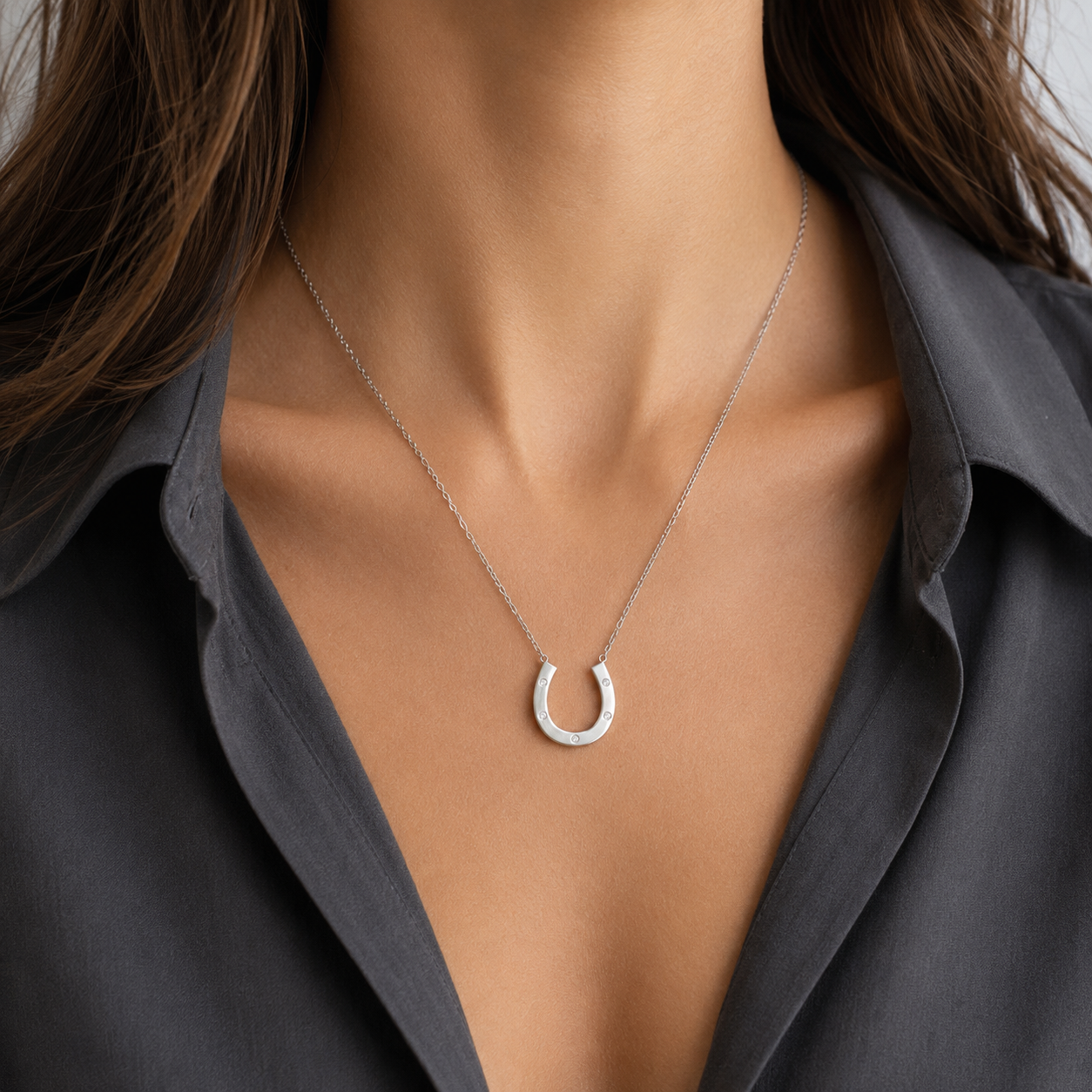 A close-up of a model wearing a delicate silver horseshoe pendant necklace with a textured, pavé-set appearance, styled with a dark gray button-down shirt.