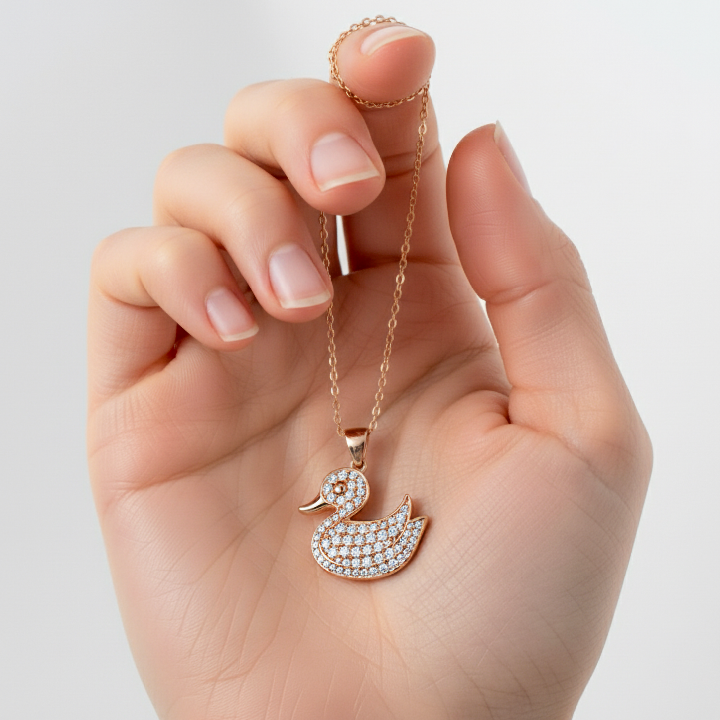 Hand holding a gold necklace with a diamond duck pendant against a light gray background