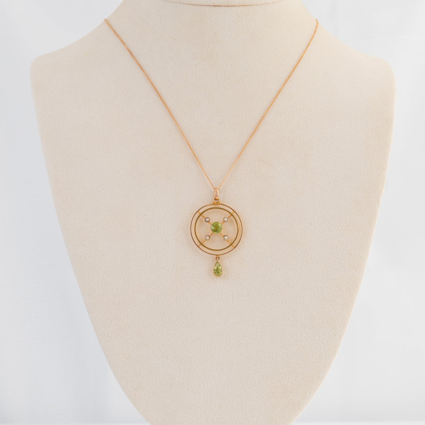 A gold pendant necklace displayed on a cream-colored jewelry bust. The necklace features a delicate gold chain and a circular pendant with a central green gemstone and small gold star-like accents, hanging from a small drop.