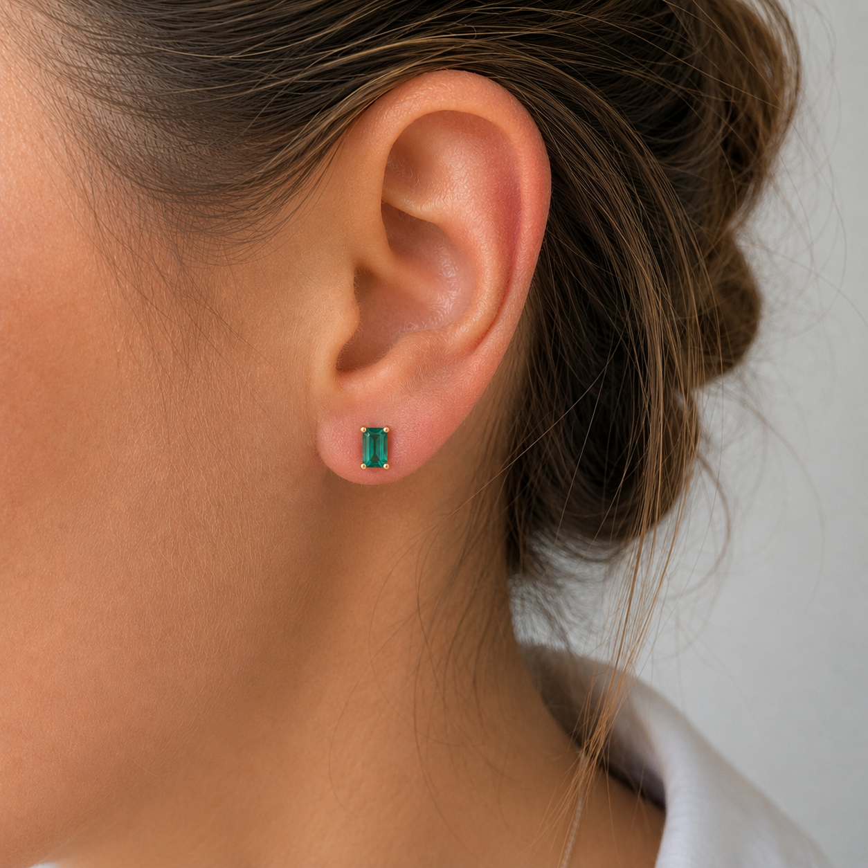 A close-up of a woman's ear wearing a small rectangular green emerald-cut stud earring in a silver setting, with her hair pulled back and a white collar visible against a light gray background.