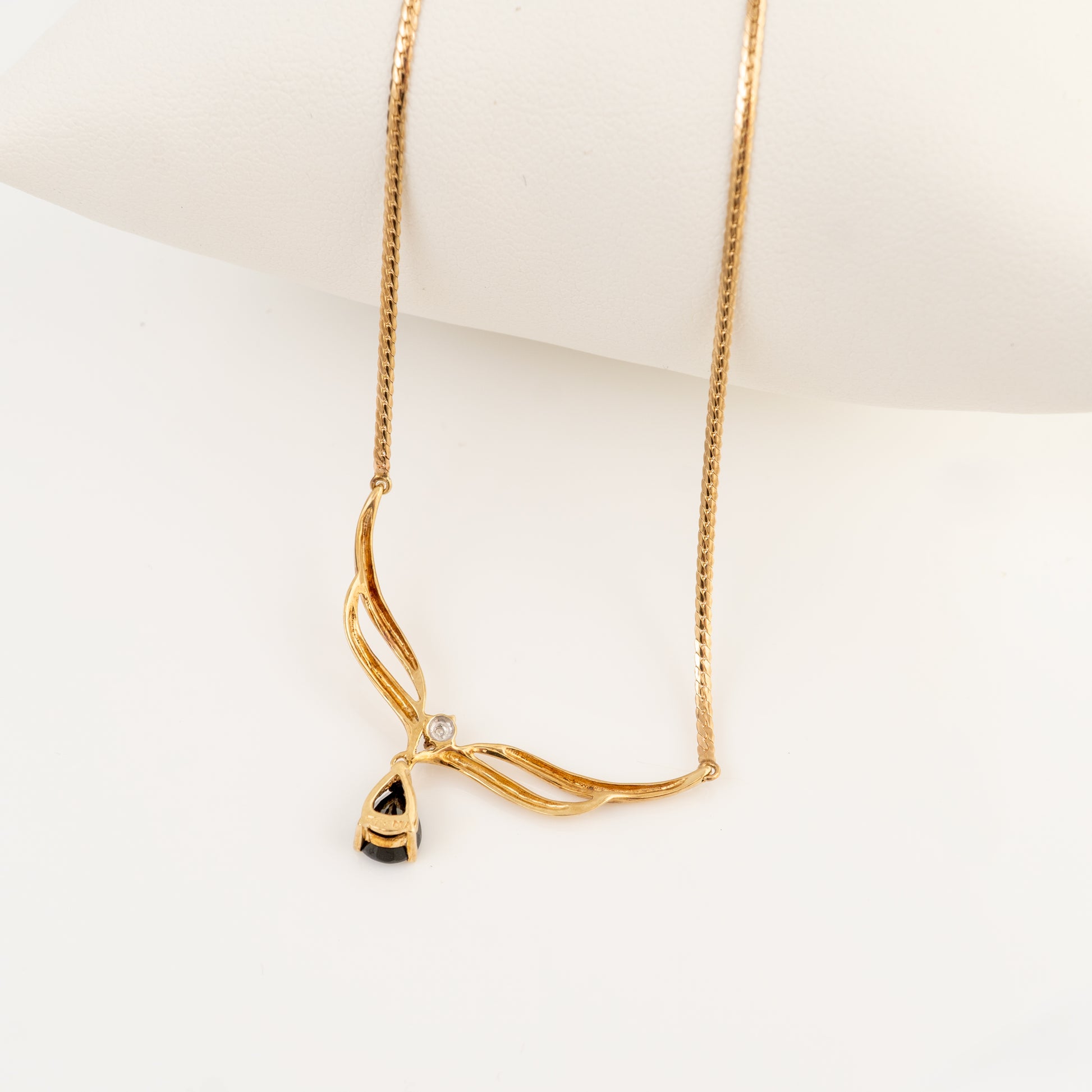 A gold necklace with a twisted, wave-like pendant design and a small clear stone, displayed on a white mannequin bust against a white background.