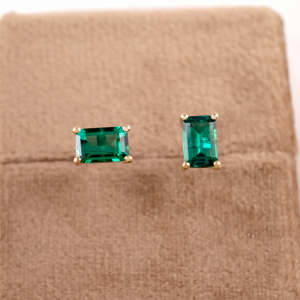A pair of rectangular cut emerald gemstone stud earrings in gold settings, displayed on a textured brown velvet jewelry stand.