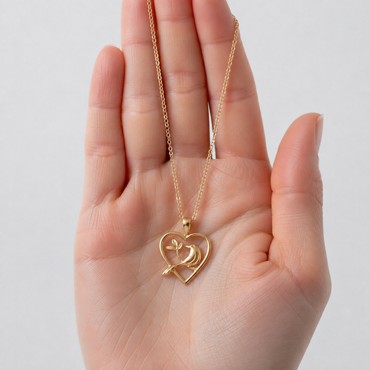 A gold heart-shaped pendant necklace with a small bird design inside, held in the palm of a hand against a light gray background.