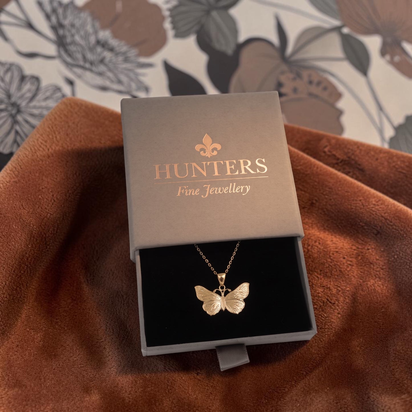 A gold butterfly pendant necklace displayed in an open Hunters Fine Jewellery gift box, resting on a brown textured fabric with a floral wallpaper background.