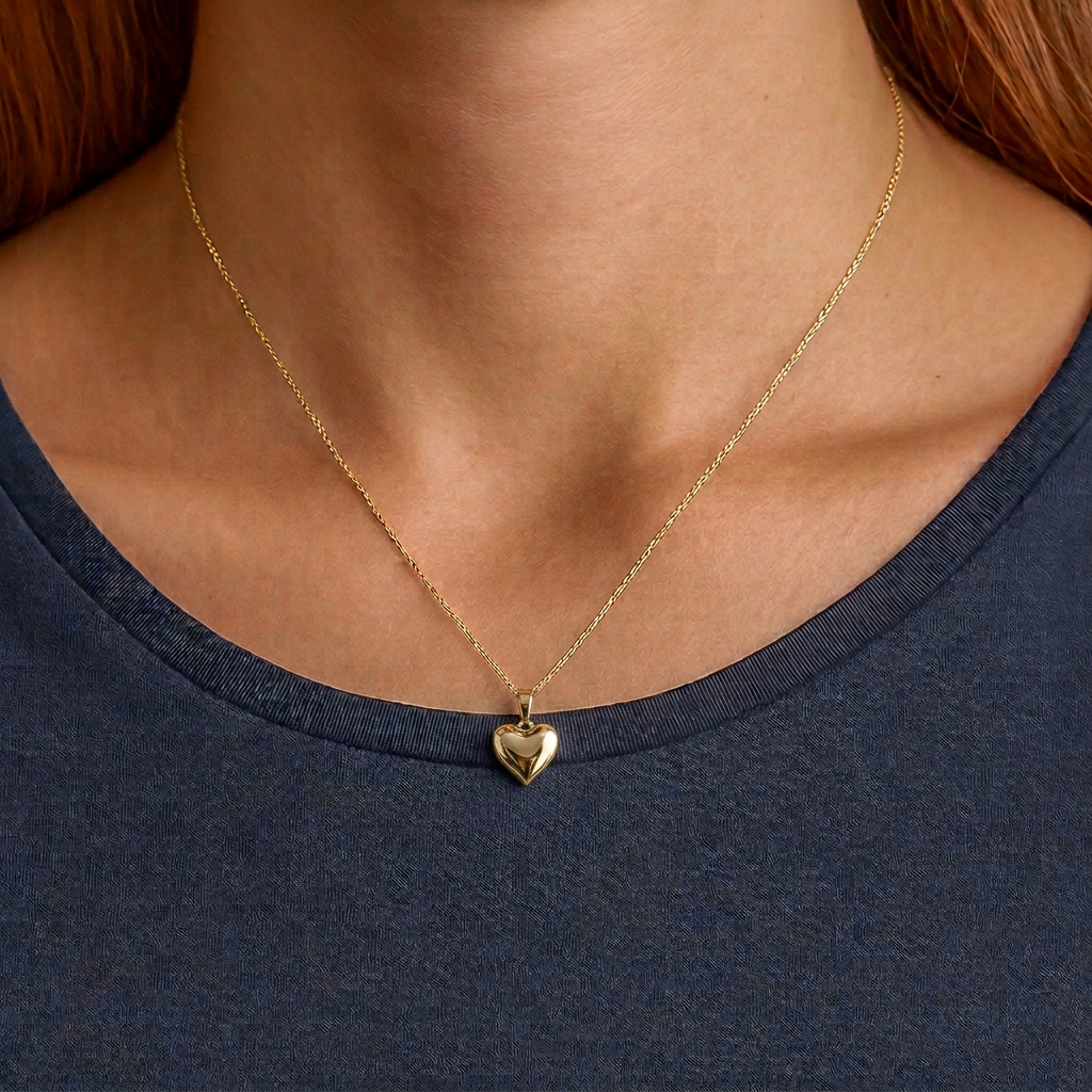A close-up of a person's neck and upper chest wearing a delicate gold chain necklace with a small heart-shaped pendant, paired with a dark heathered blue-gray t-shirt.