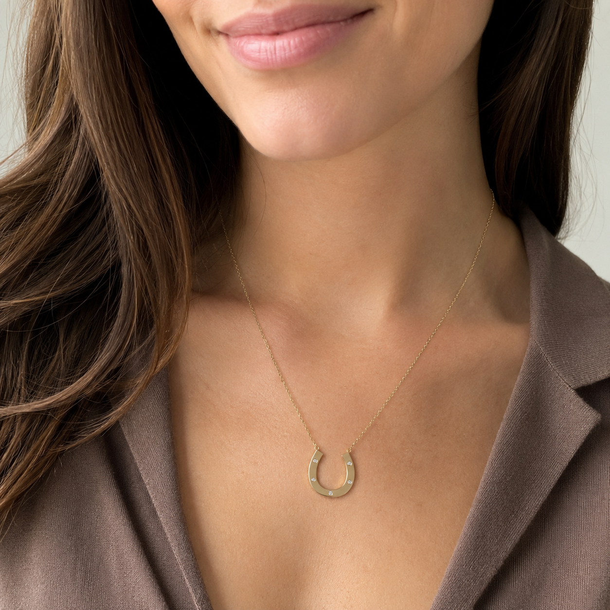 A close-up of a woman wearing a delicate gold horseshoe pendant necklace, styled with a brown blazer.