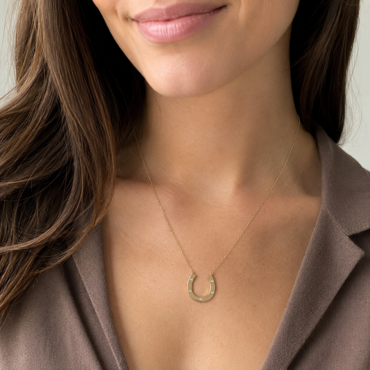 A close-up of a woman wearing a delicate gold horseshoe pendant necklace, styled with a brown blazer.