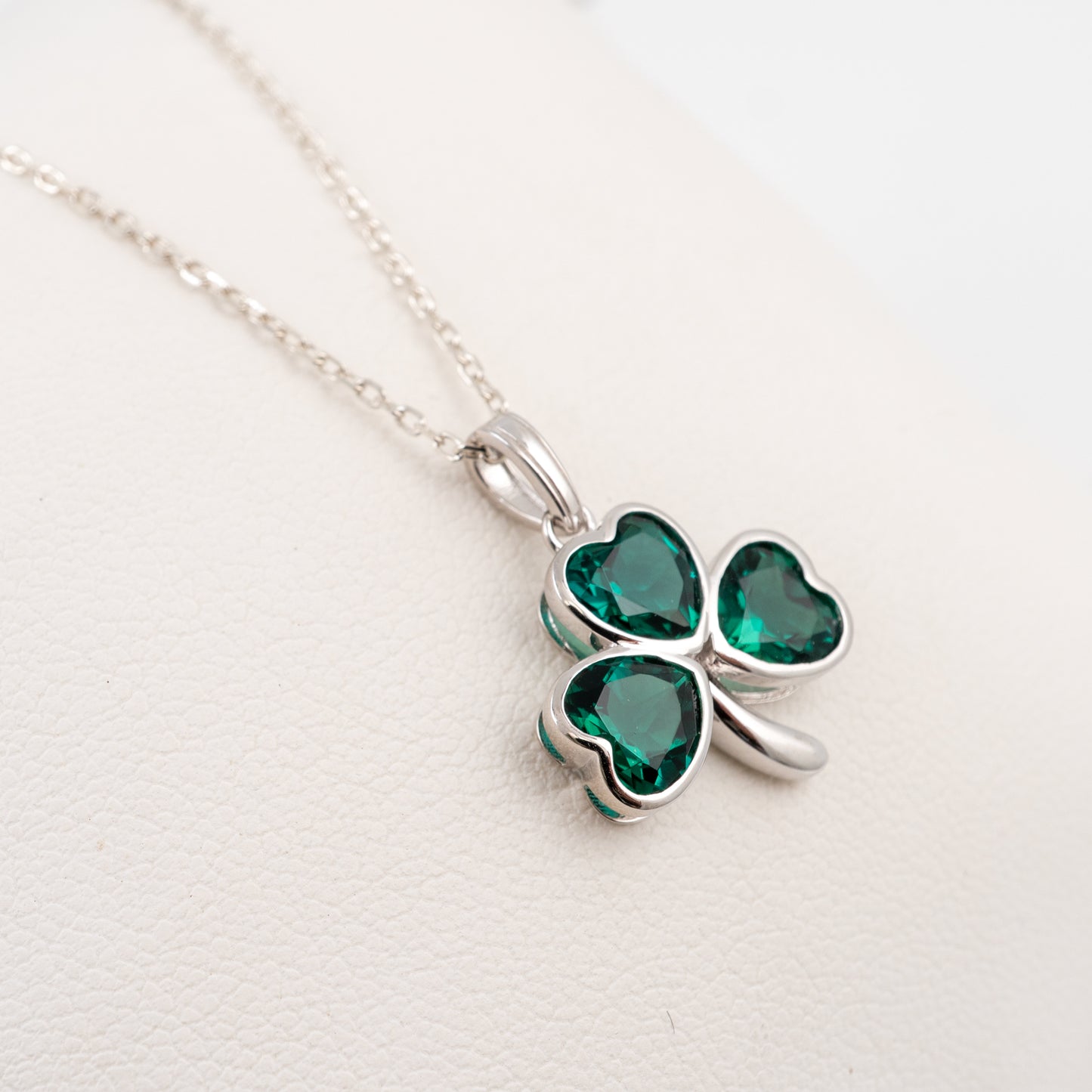 A silver shamrock pendant necklace with three heart-shaped green gemstones, displayed on a textured white surface.