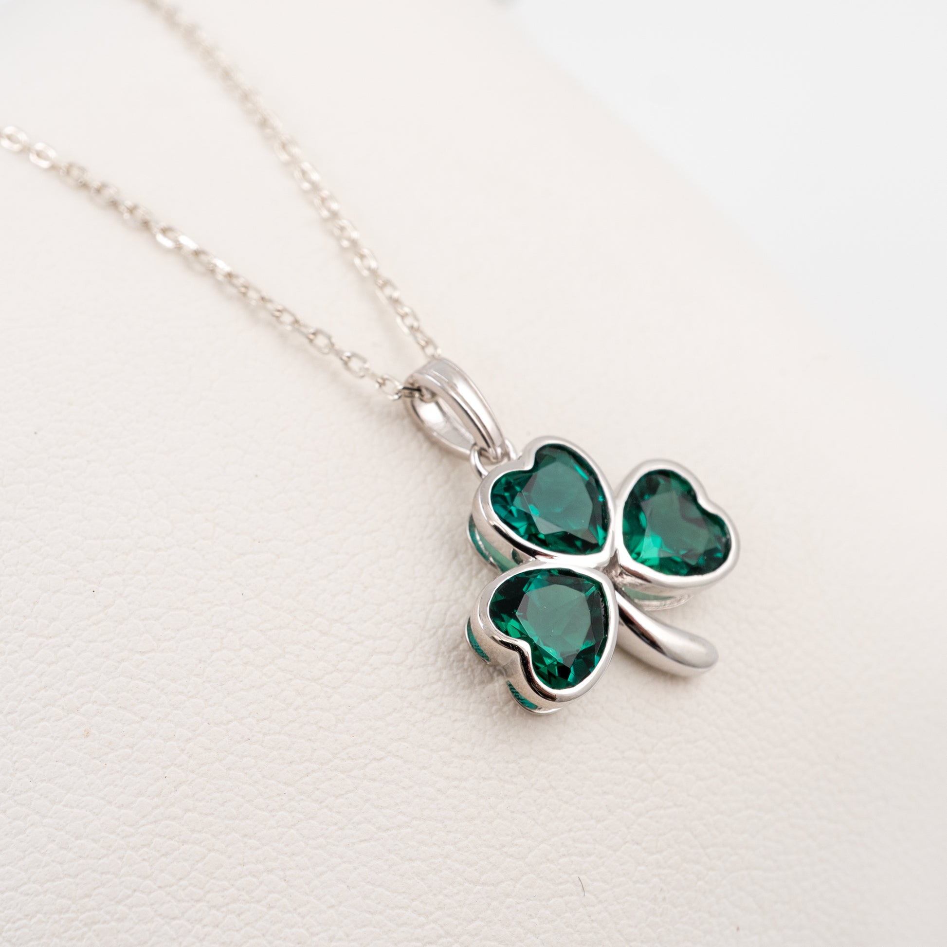 A silver shamrock pendant necklace with three heart-shaped green gemstones, displayed on a textured white surface.