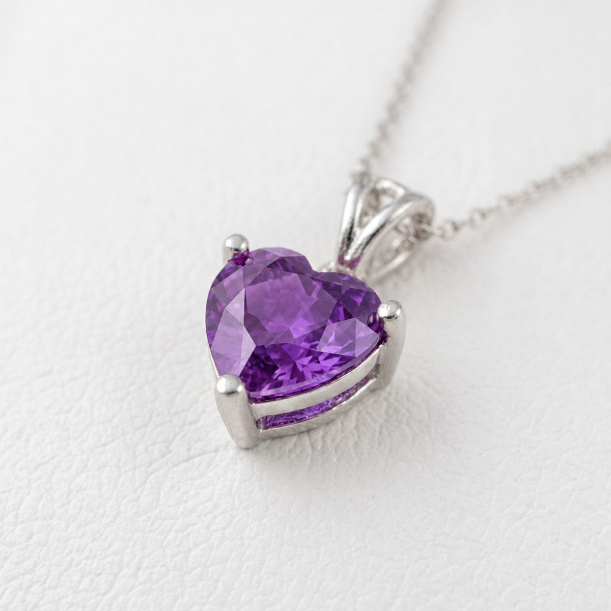A heart-shaped amethyst pendant in a silver setting on a delicate silver chain, photographed on a textured white surface.