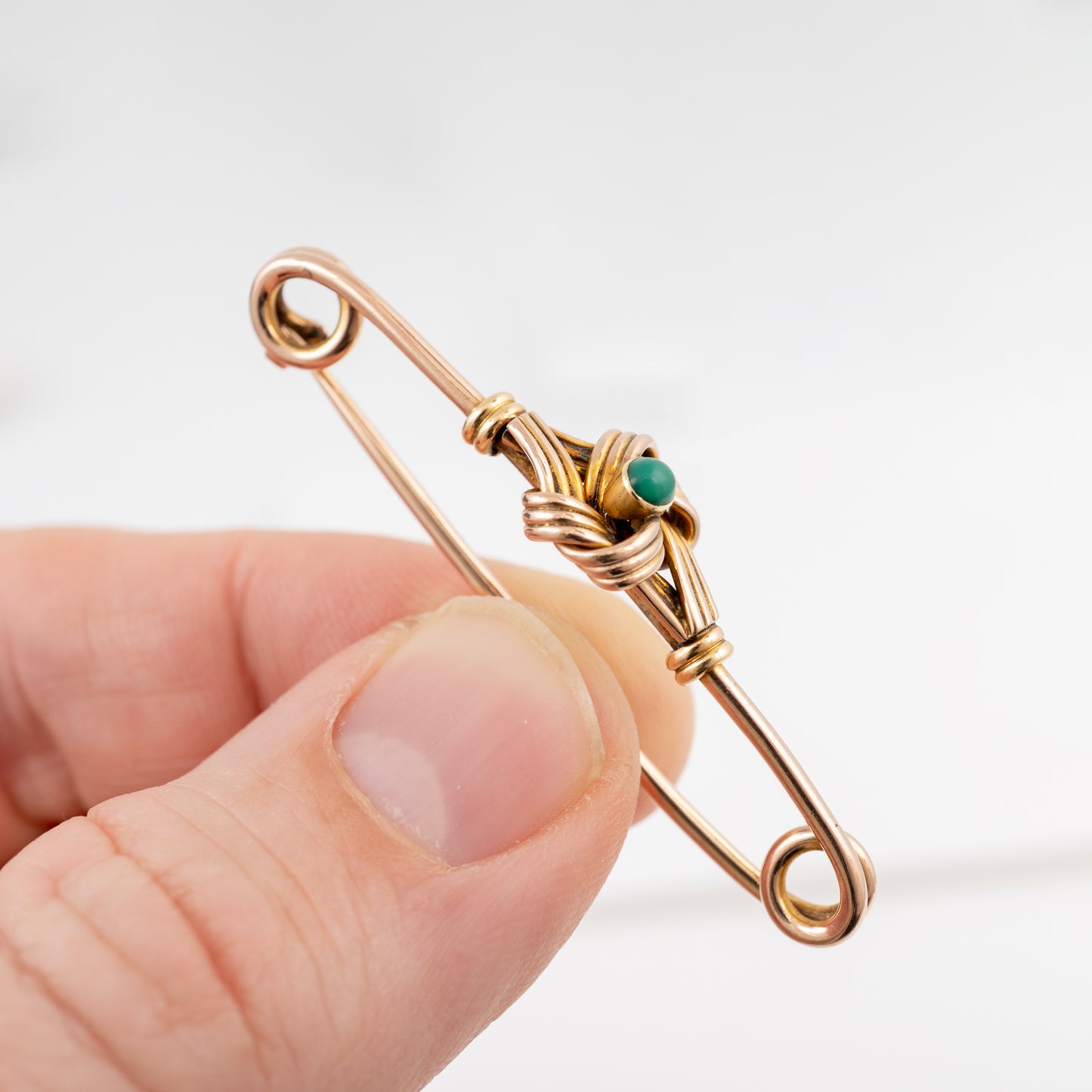A close-up of a hand holding a small safety pin brooch made of copper-toned wire with a tiny green stone at the center, against a plain light background.