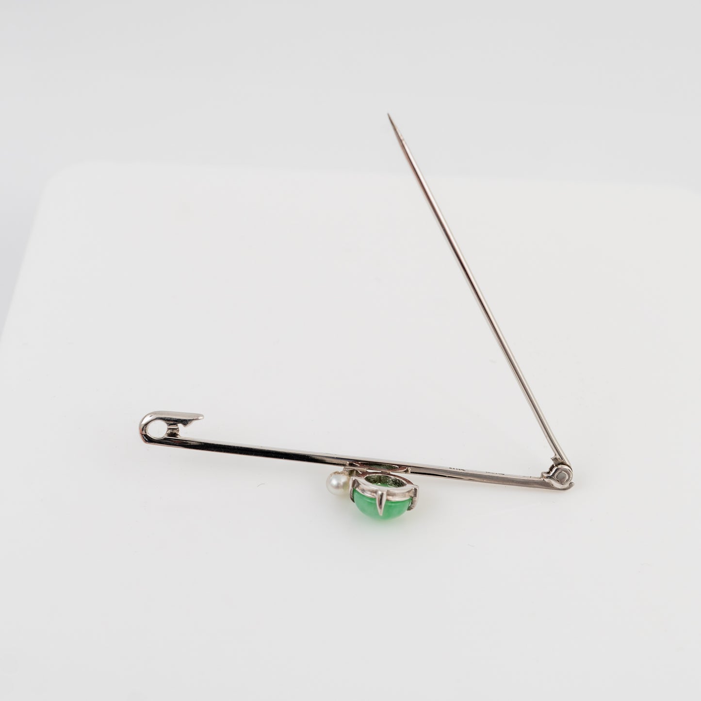 A white gold pin styled as a brooch, featuring a small green faceted gemstone in a bezel setting at the center, displayed on a clean white background.