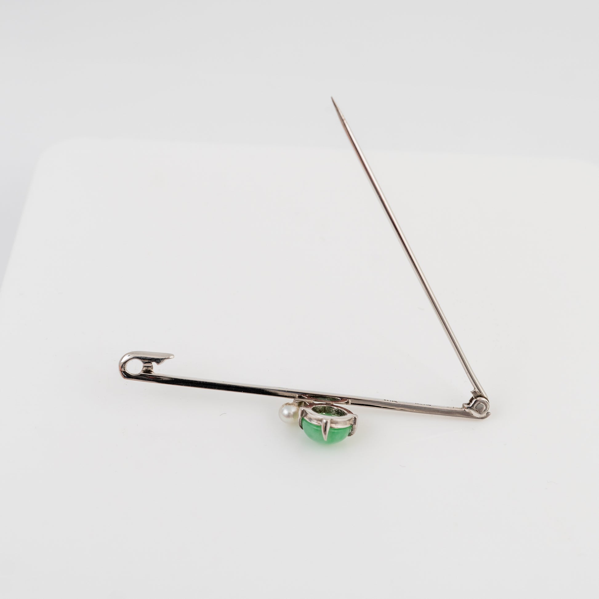 A white gold pin styled as a brooch, featuring a small green faceted gemstone in a bezel setting at the center, displayed on a clean white background.