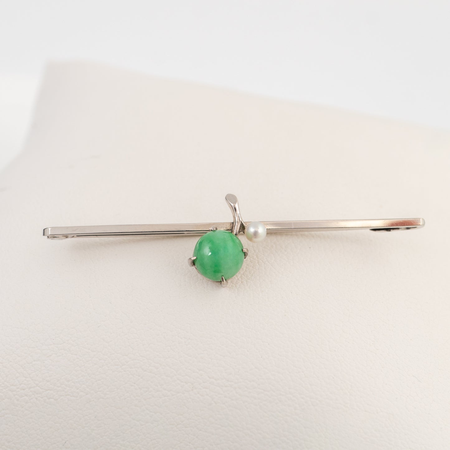 A white gold brooch pin with a round green jadeite gemstone and a small white pearl, displayed on a cream-coloured jewelry stand.