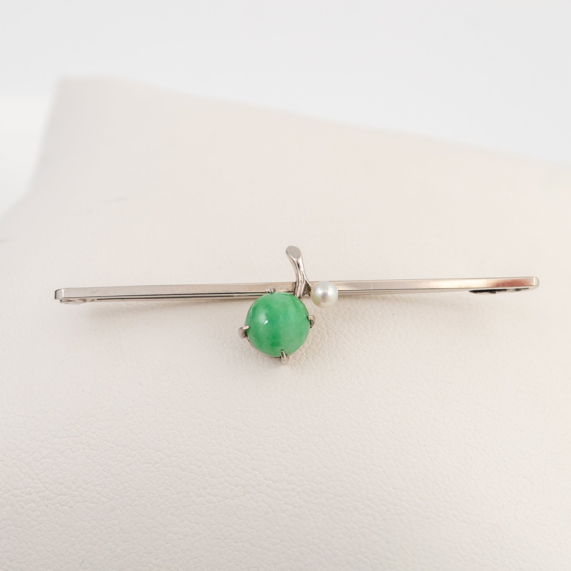 A white gold brooch pin with a round green jadeite gemstone and a small white pearl, displayed on a cream-coloured jewelry stand.