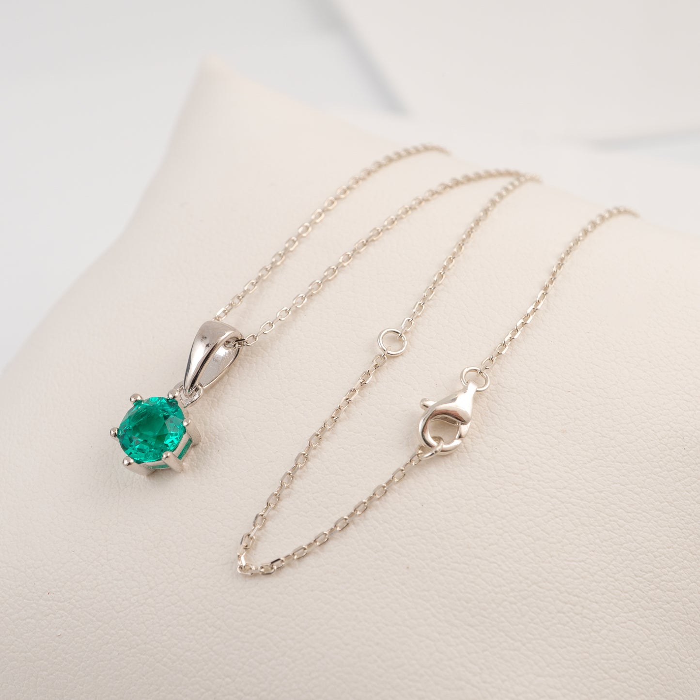 A silver pendant necklace with a round-cut green emerald gemstone, displayed on a cream-colored jewelry stand.