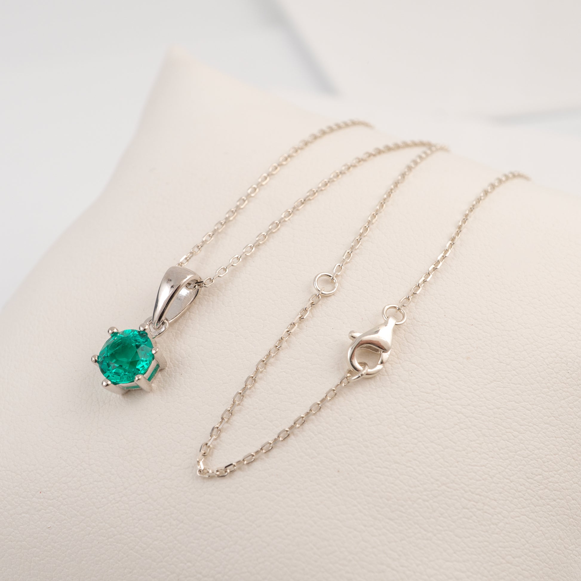 A silver pendant necklace with a round-cut green emerald gemstone, displayed on a cream-colored jewelry stand.