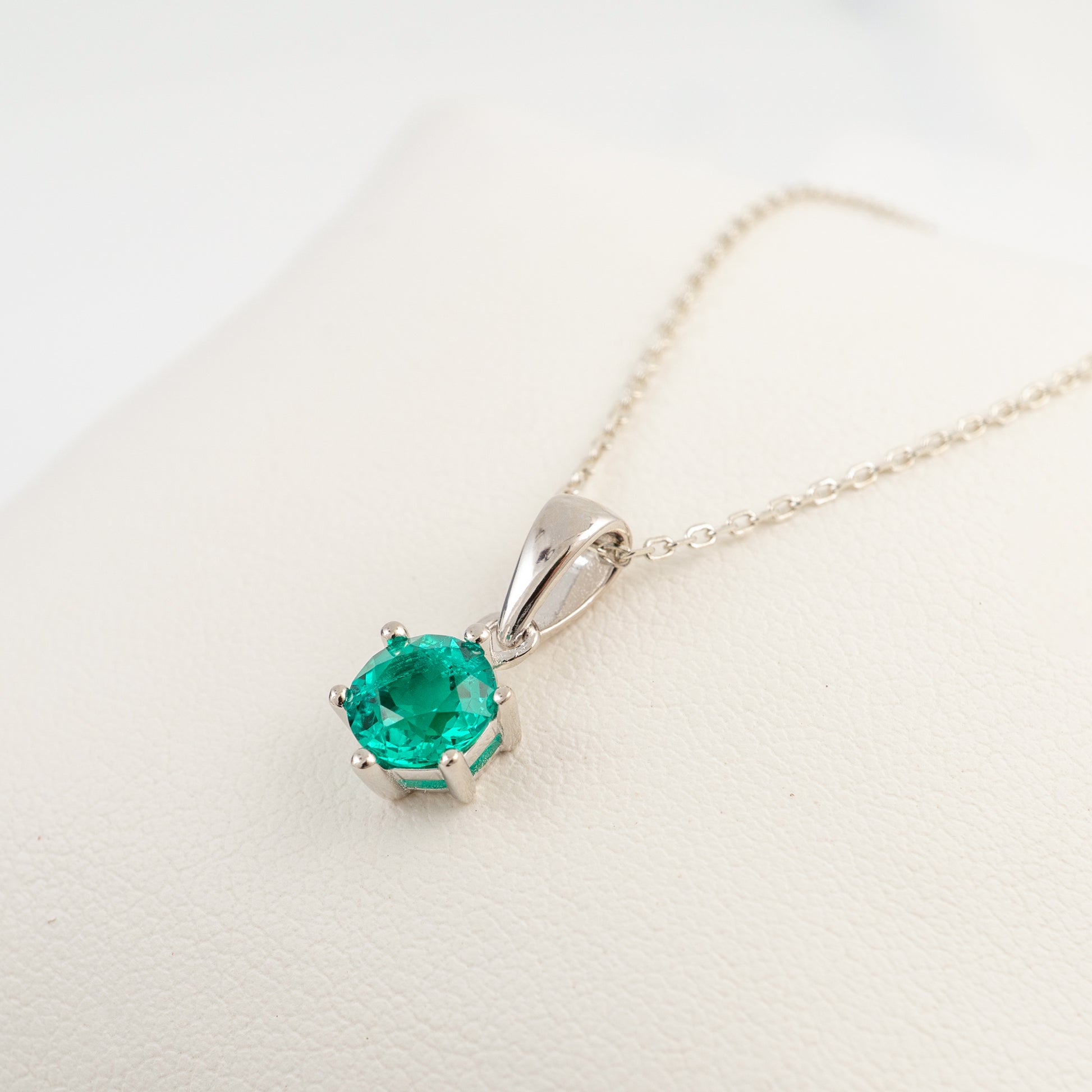 A silver pendant necklace featuring a round-cut green emerald gemstone in a prong setting, resting on a textured white surface.