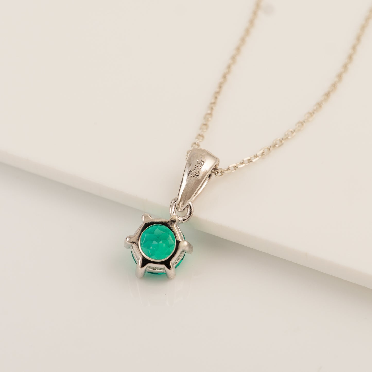A silver pendant necklace featuring a round green gemstone in a prong setting, resting on a clean white surface.