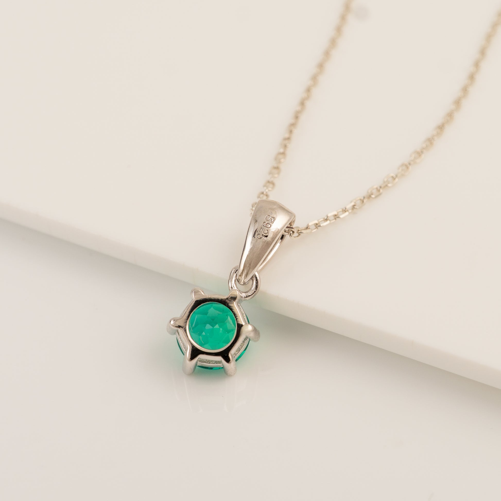 A silver pendant necklace featuring a round green gemstone in a prong setting, resting on a clean white surface.