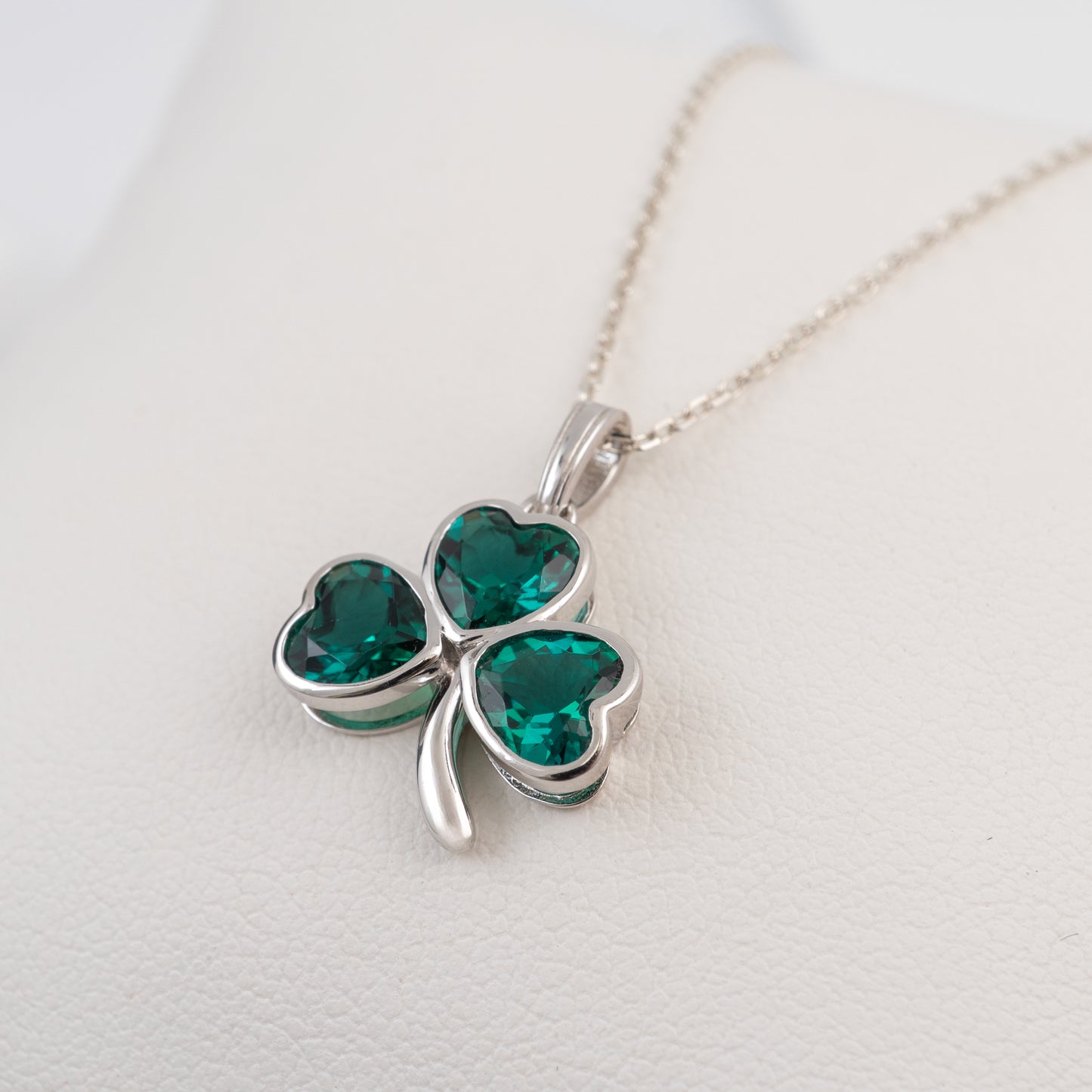 A silver shamrock pendant necklace with three heart-shaped emerald green crystals, displayed on a textured white surface.