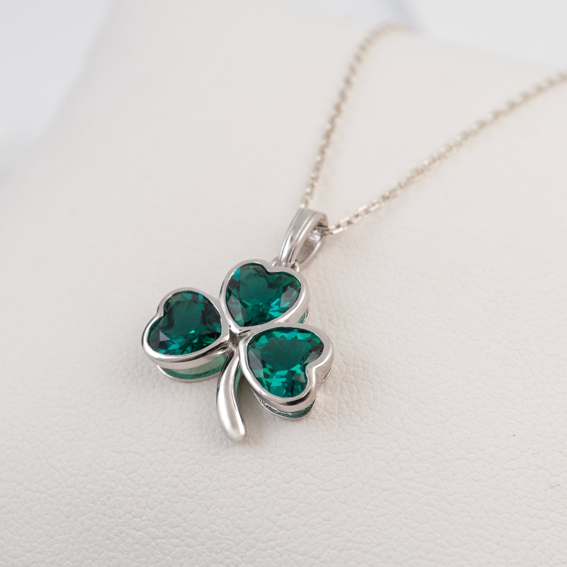 A silver shamrock pendant necklace with three heart-shaped emerald green crystals, displayed on a textured white surface.