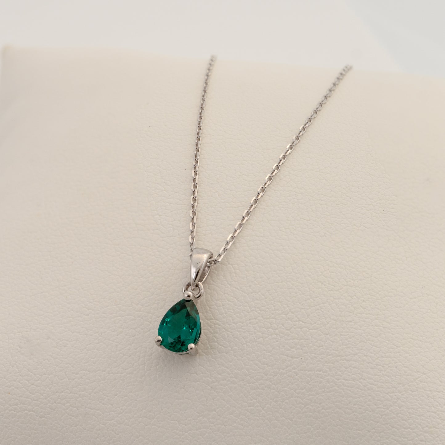 Teardrop green lab emerald gemstone pendant on a silver chain against a beige background