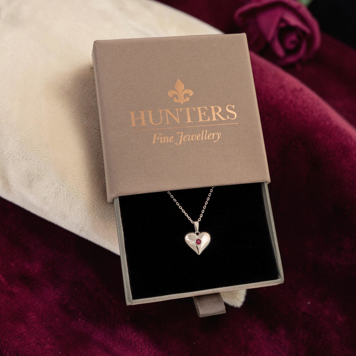 A silver heart pendant necklace with a small lab grown ruby gemstone lies in an open taupe jewelry box. The box lid is printed with the brand name 'HUNTERS Fine Jewellery' in gold.
