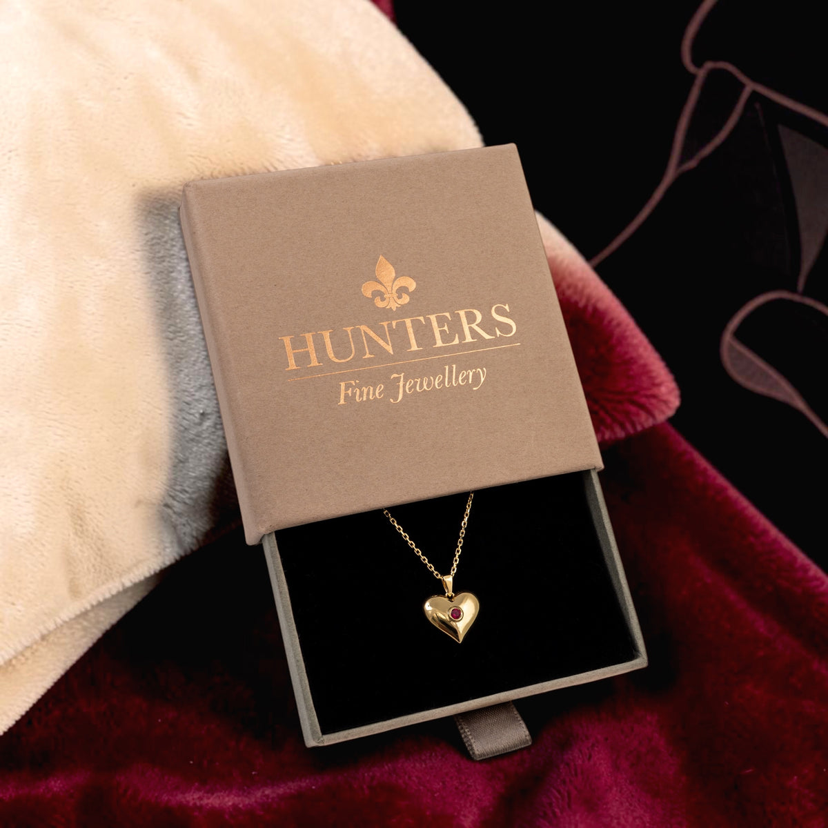 A gold heart pendant necklace displayed in an open Hunter's Fine Jewellery gift box, set against a background of cream faux fur and deep burgundy velvet.