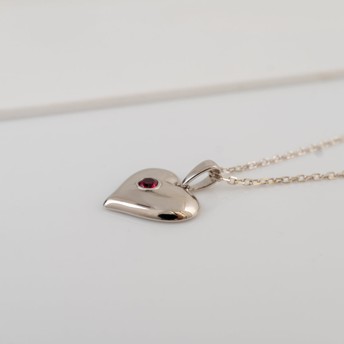 A polished silver heart pendant necklace with a small red gemstone accent, resting on a light gray surface.