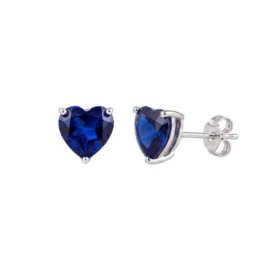 A pair of heart-shaped royal blue sapphire stud earrings in silver settings, with one earring facing forward and the other showing the post and backing, on a plain white background.