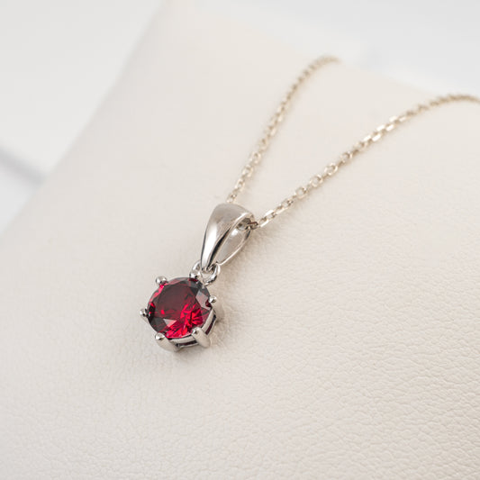 A silver pendant necklace with a round red gemstone, resting on a textured cream-colored surface.