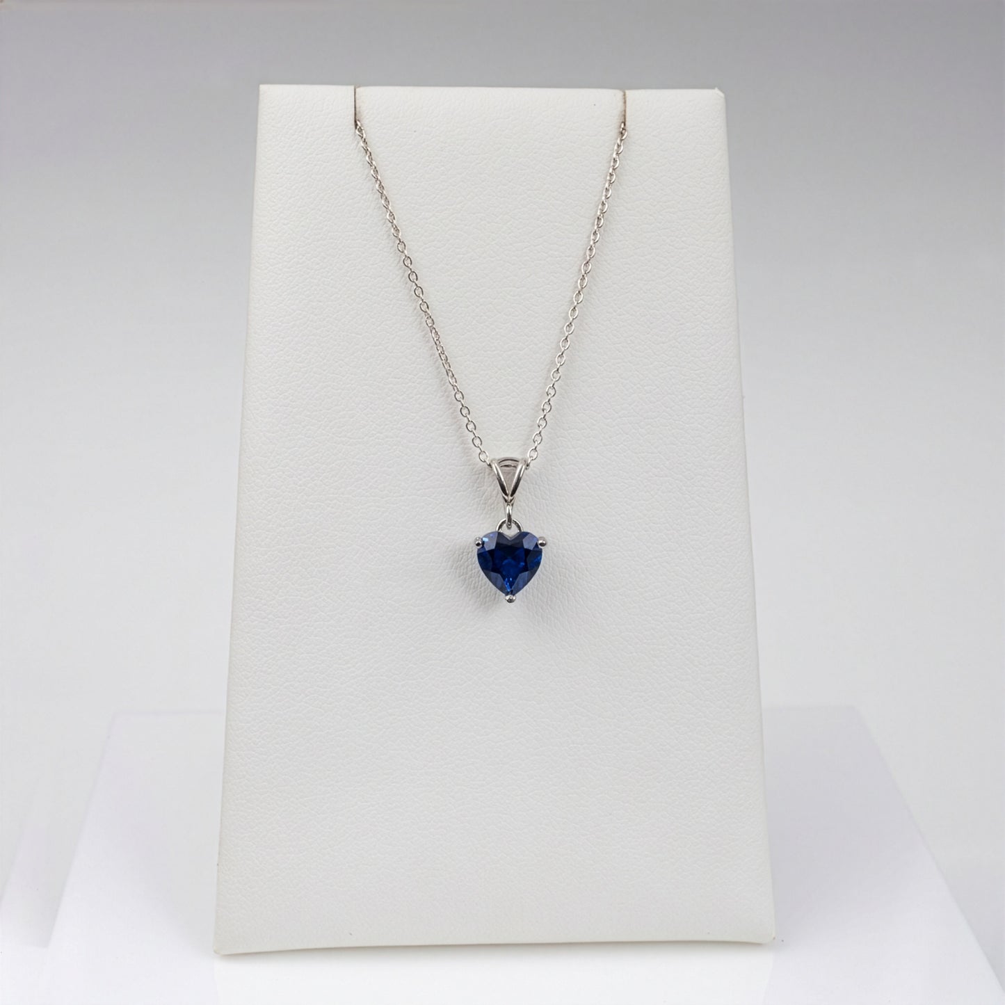A silver pendant necklace with a heart-shaped blue lab sapphire gemstone, displayed on a white jewelry stand against a light gray background.
